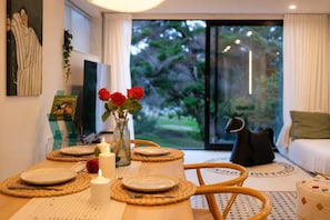 A dining area with a table set for two, a vase with red roses, and a view of a garden through a glass door. - Stylish House with Great City Views (Auckland)