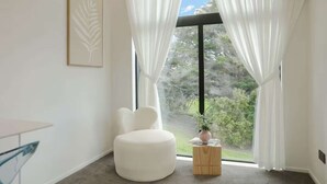 Een moderne kamer met een groot raam, een witte stoel, een klein houten bijzettafeltje en een lijstwerk kunstwerk aan de muur.