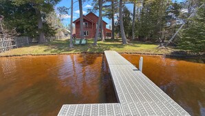 Un muelle de madera que se adentra en un cuerpo de agua, con una casa roja al fondo rodeada de árboles.