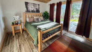 Una cama de madera con una colcha verde, una mesita de noche con una lámpara y vistas al exterior a través de amplios ventanales.