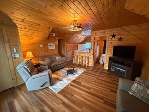 A cozy log cabin interior with a sofa, armchair, TV, and a small kitchen area.