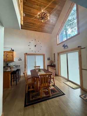 A dining area with a wooden table and chairs, a chandelier, a wall clock, and a large window.
