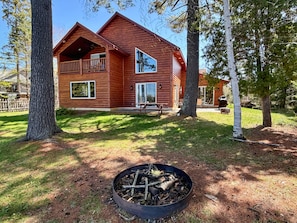 A two-story log cabin with a balcony, a fire pit, and a grassy yard.
