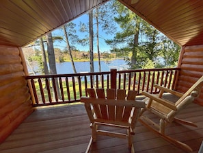 A wooden cabin with a deck, rocking chairs, and a view of a lake and trees.