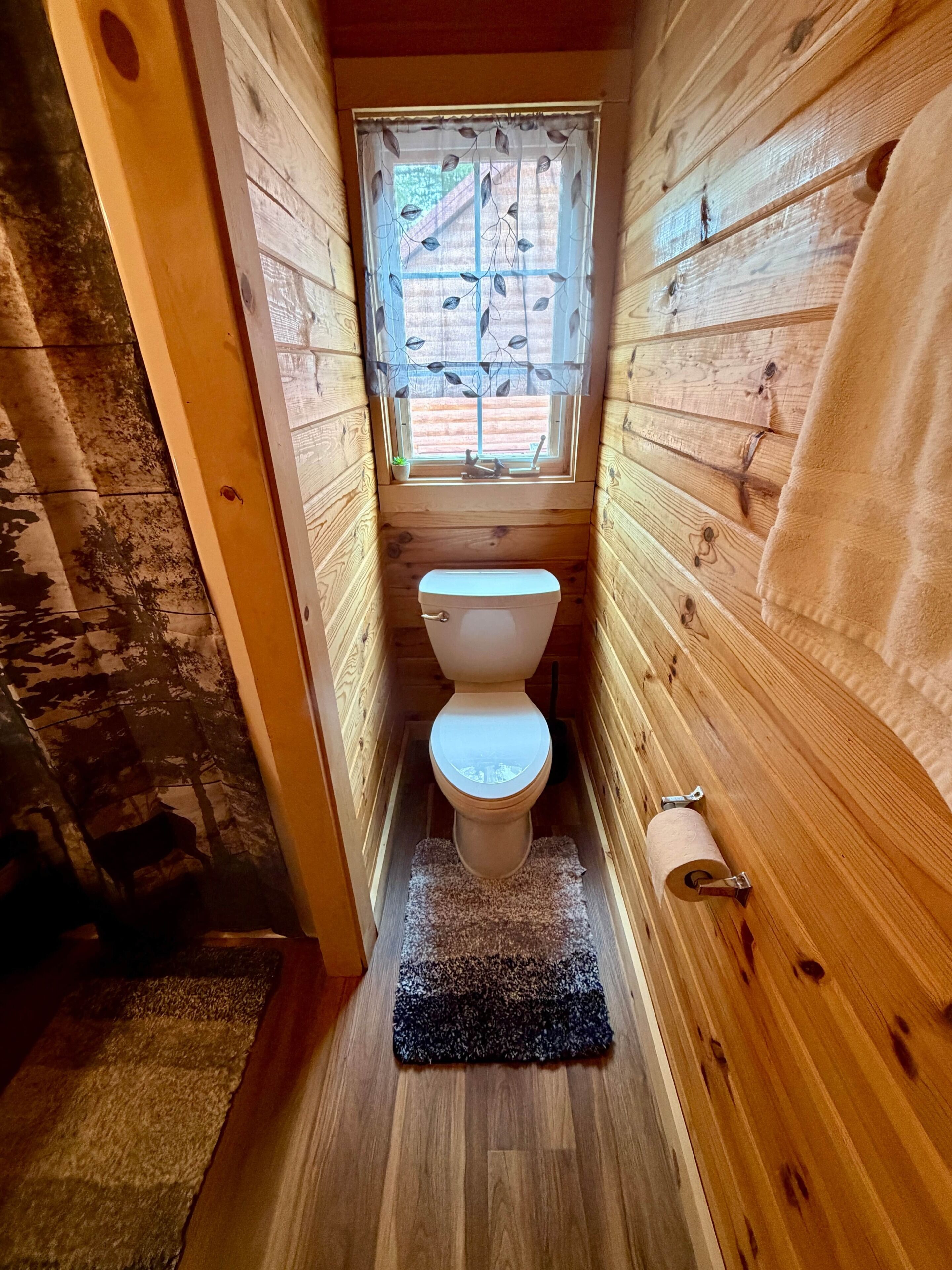 Een smalle houten badkamer met toilet, een raam met gordijn met bladmotief, en een houten vloer.