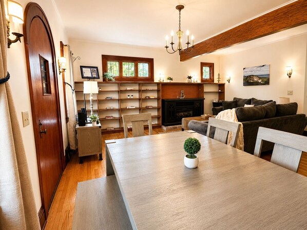 A dining area with a wooden table, chairs, a chandelier, a fireplace, and a bookshelf.