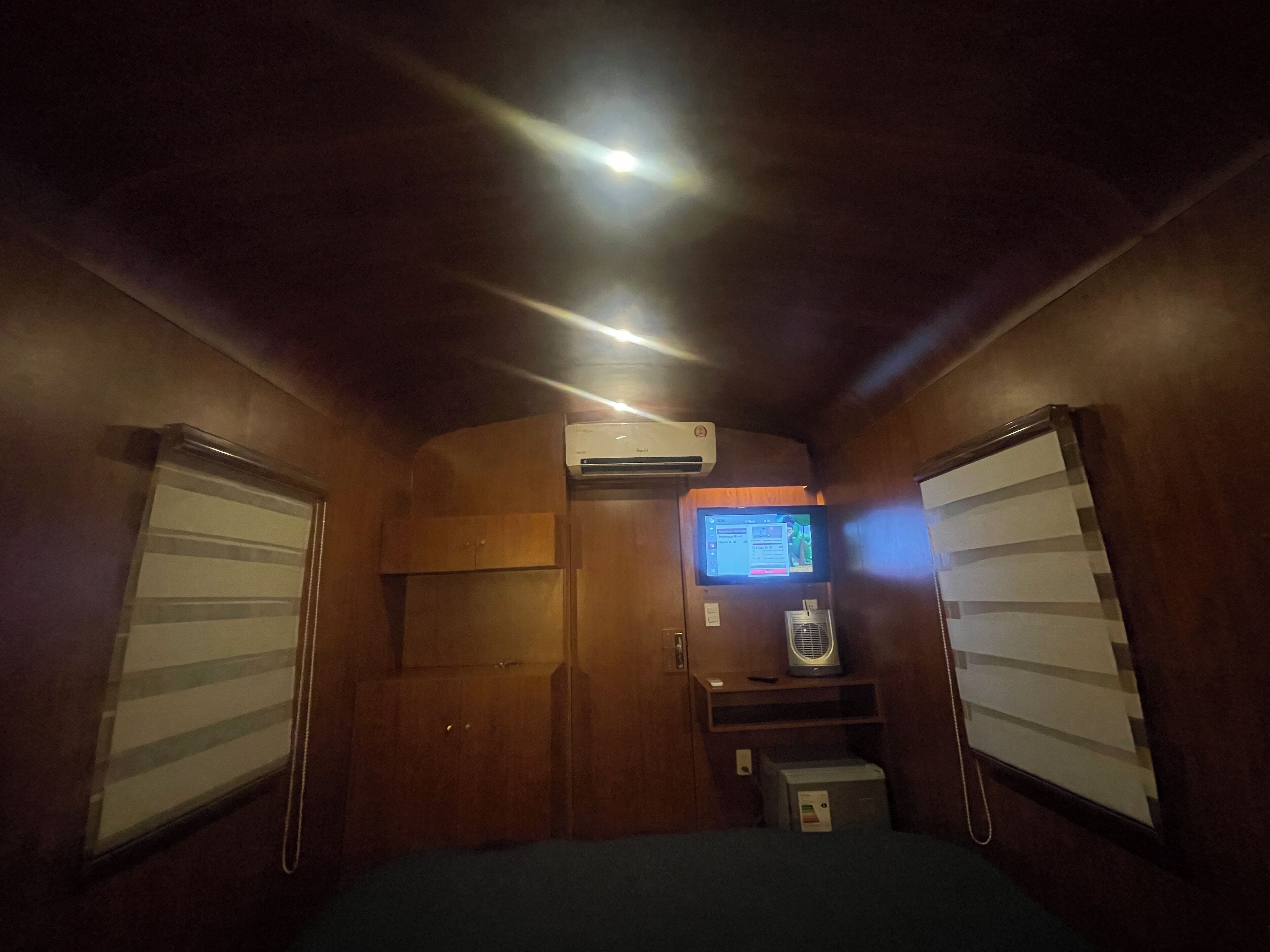 A compact room with wooden walls, a small bed, an air conditioning unit, a television, and a shelf with a fan.