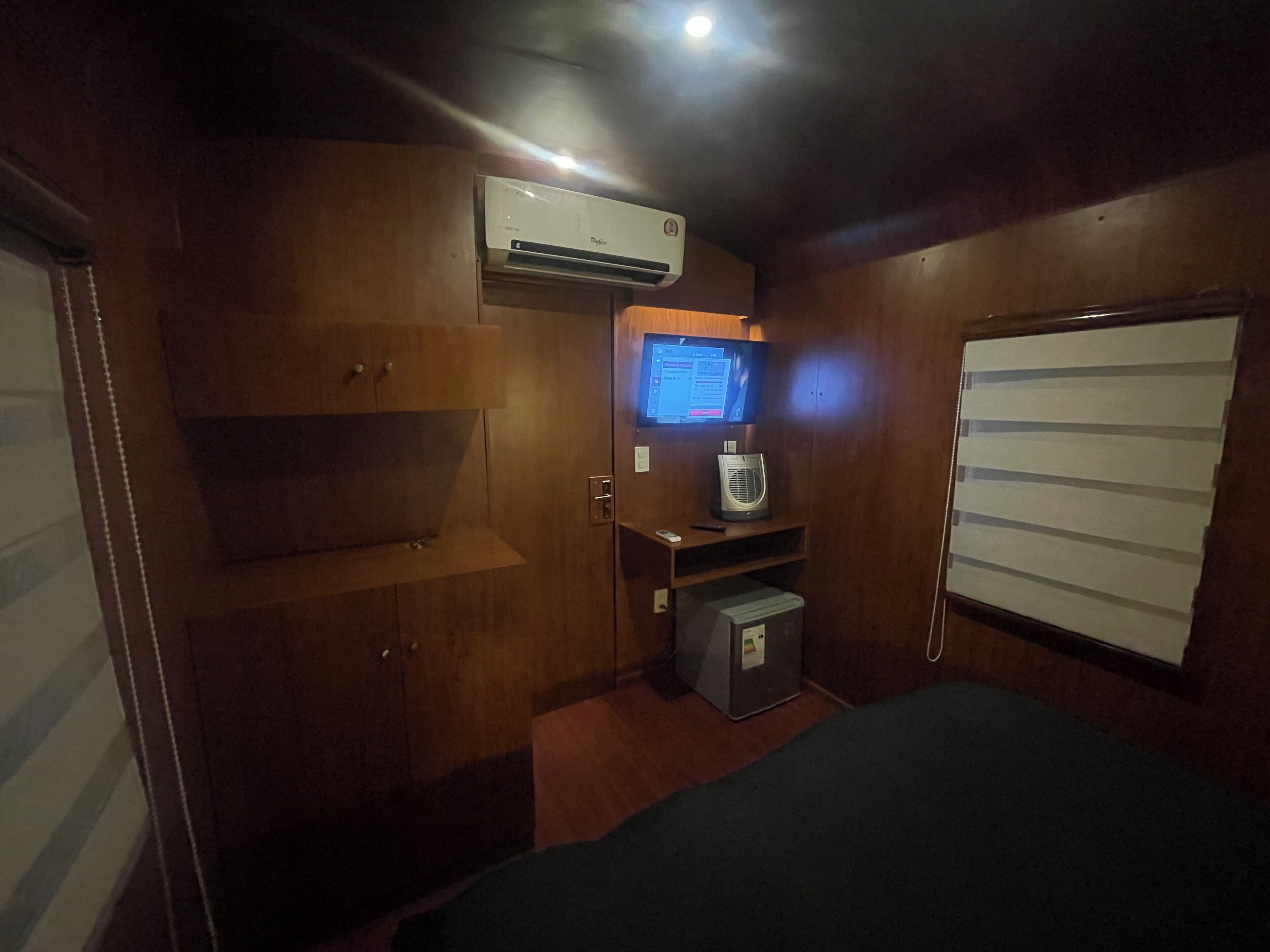 A compact room with a bed, wooden cabinets, a small desk with a computer, and a wall-mounted air conditioner.
