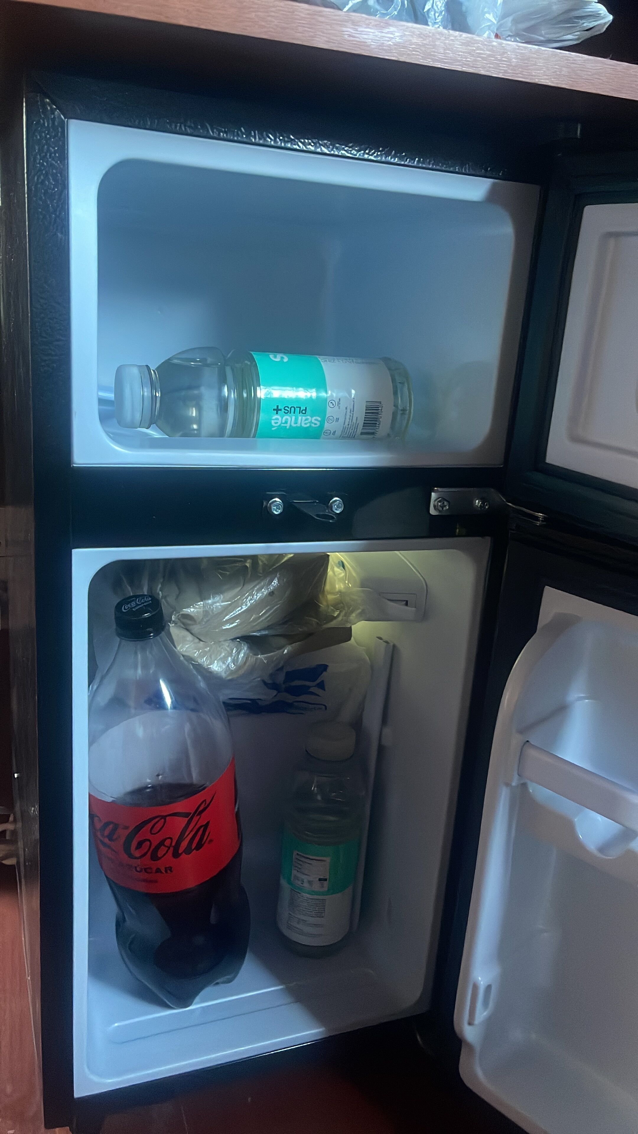 A refrigerator with two shelves, one containing a Coca-Cola bottle and a bottle of water, and the other with a plastic bag and a bottle of dish soap.