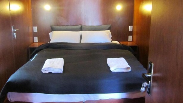 A hotel room with a wooden headboard, two white pillows, a black bedspread, and two folded white towels on the bed. - Onkel Inn Wagon Sleepbox (Uyuni)