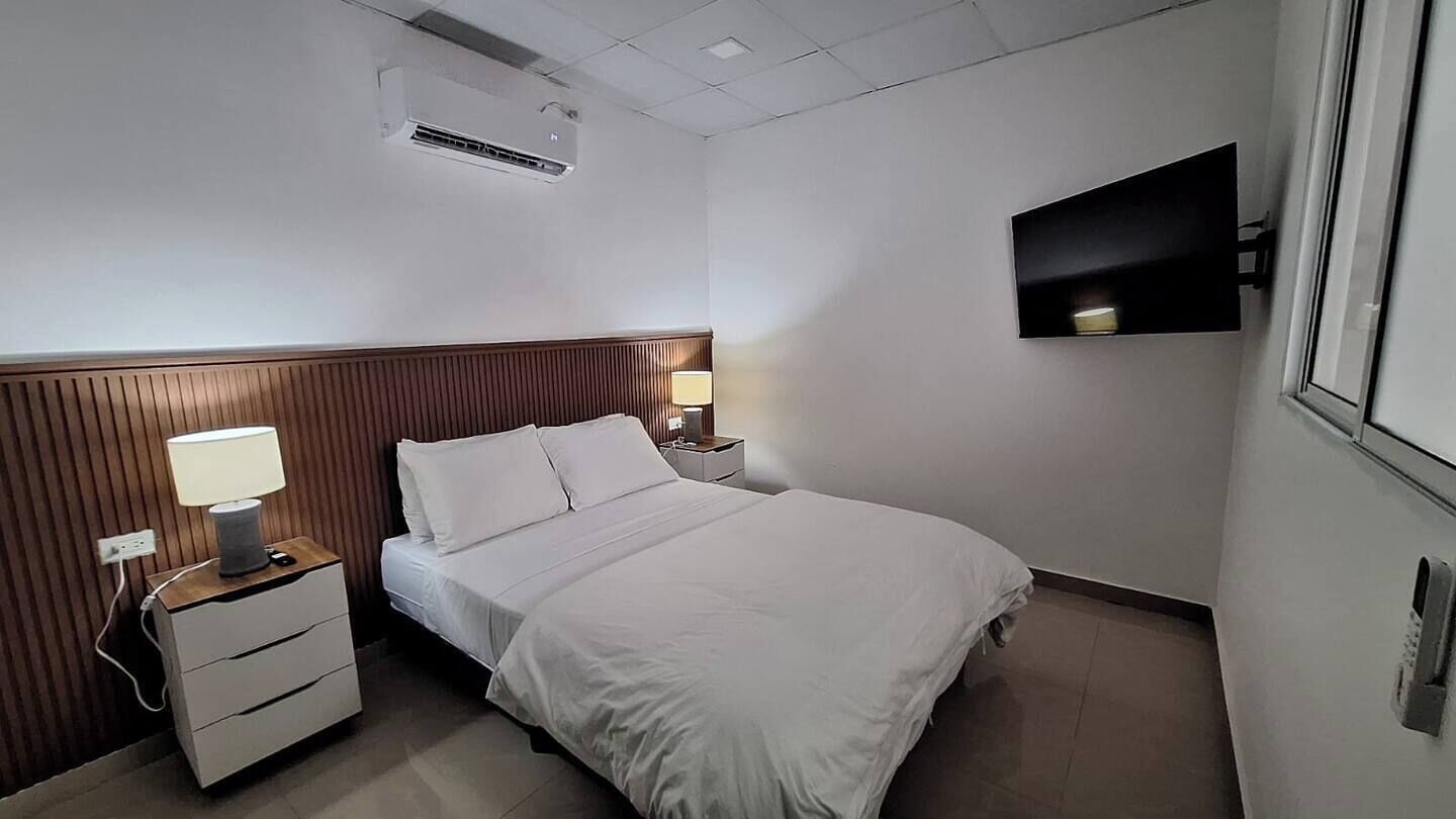 A hotel room with a bed, a nightstand with a lamp, a wall-mounted TV, and an air conditioning unit.