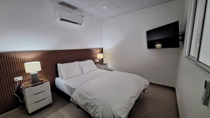 A hotel room with a bed, a nightstand with a lamp, a wall-mounted TV, and an air conditioning unit. - apto-wifi6-tvstreaming (Panamá)