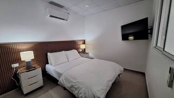 A hotel room with a bed, a nightstand with a lamp, a wall-mounted TV, and an air conditioning unit.
