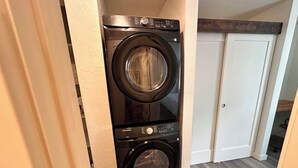 Une machine à laver et un sèche-linge superposés sont installés dans un couloir, sur un plancher en bois, avec une porte blanche en arrière-plan.