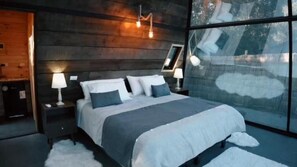A modern bedroom with a large bed, wooden headboard, and a view of the outdoors through large windows. - Cabañas Boutique Aires del toro (Curacaví)