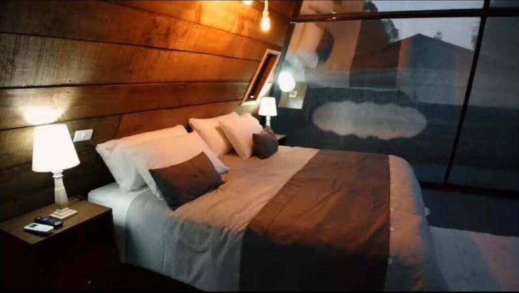A bedroom with a wooden headboard, a bed with white bedding, two bedside lamps, and a large window with a view of the outside.