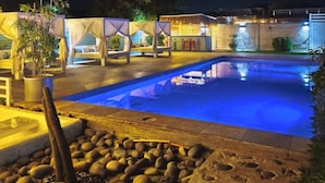 Ein Schwimmbeckenbereich bei Nacht mit Pool, Liegestühlen und einem Kaktus im Vordergrund.