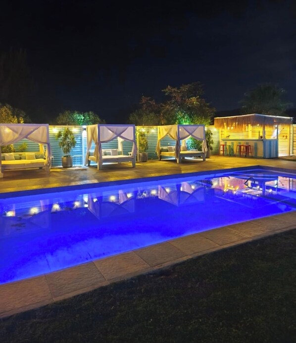 Une piscine éclairée de lumières bleues, un bar avec des tabourets et un espace extérieur avec des auvents.