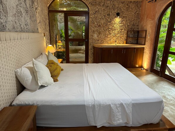 Une chambre à coucher avec une tête de lit ornée d’un motif en briques, un lit recouvert de linge de maison blanc, une table de chevet en bois, une lampe de chevet et une vue sur un mur de pierre et la verdure à l’extérieur.