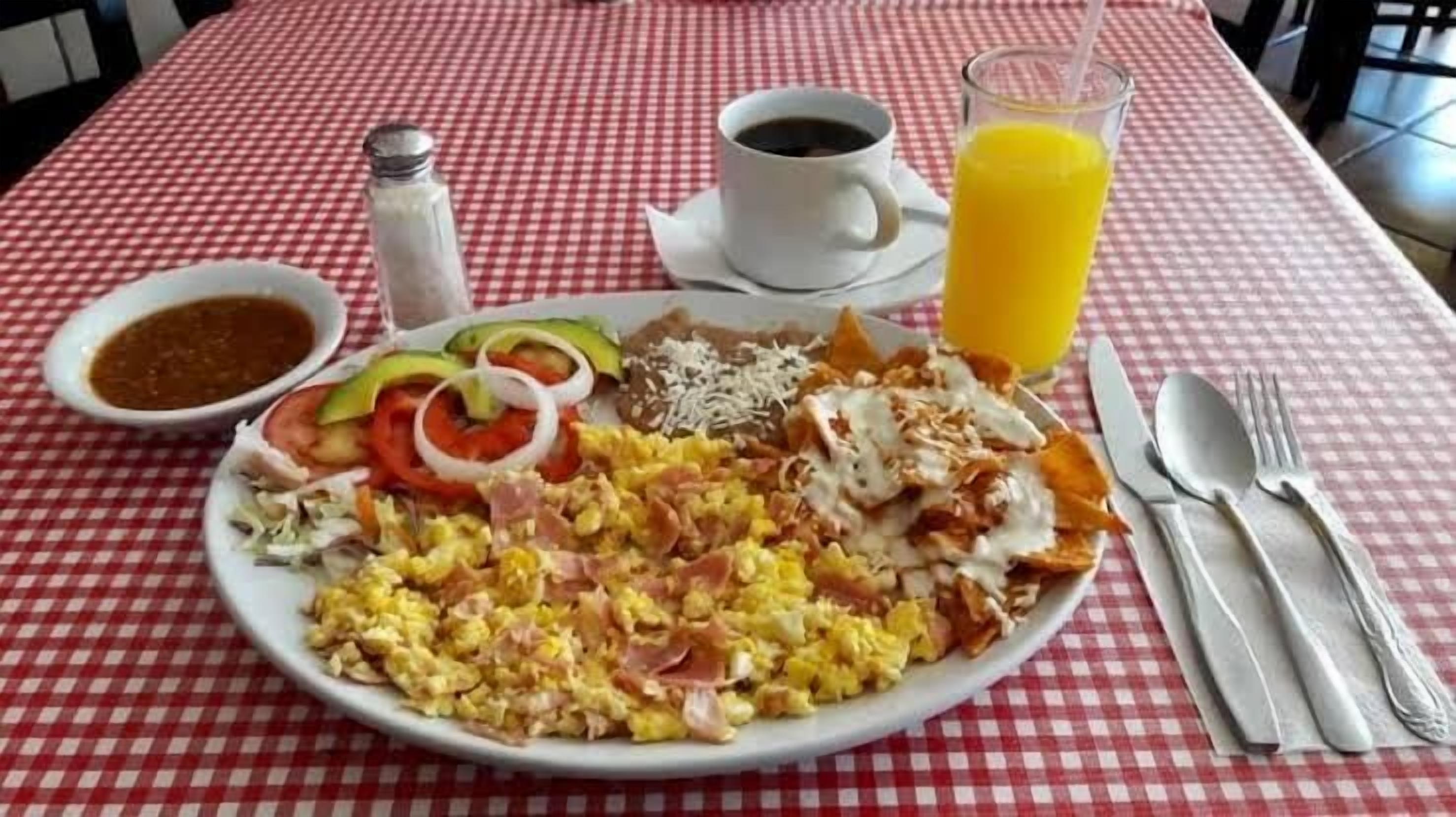 Une assiette de petit-déjeuner contenant des œufs brouillés, du bacon, du fromage et des légumes, une tasse de café et un verre de jus d’orange, le tout posé sur une nappe à carreaux.