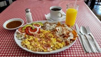 Une assiette de petit-déjeuner contenant des œufs brouillés, du bacon, du fromage et des légumes, une tasse de café et un verre de jus d’orange, le tout posé sur une nappe à carreaux.