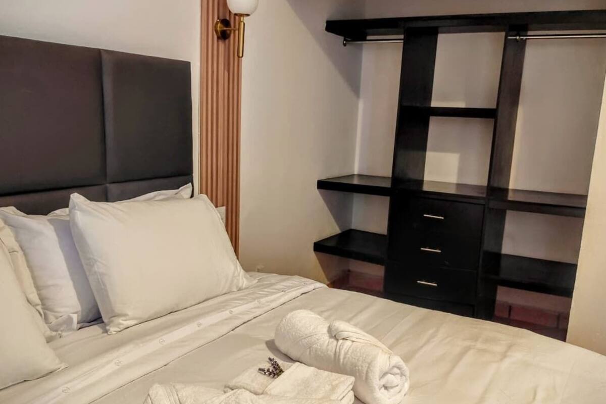 A hotel room with a bed, two rolled towels, a black wardrobe, and a wooden panel wall.