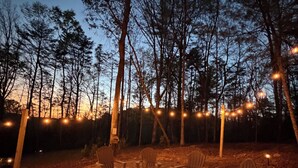 Un espacio exterior con sillas Adirondack y una fogata, rodeado de árboles y luces colgantes.