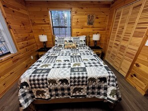 Ein Schlafzimmer einer Holzhütte mit einer karierten Bettdecke mit Bärmotiven, zwei Nachttischlampen, einem Fenster mit Jalousien und einer Holztür.