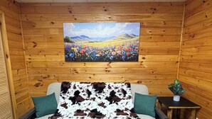Interior de una cabaña de madera con sofá, almohadas, un cuadro de un campo y una planta pequeña.