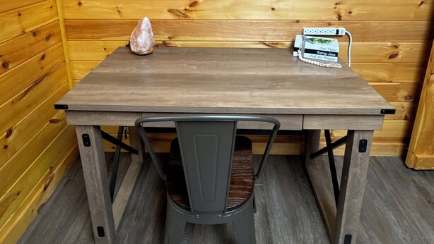 Interior de una cabaña de madera con una mesa de comedor, dos sillas, un cuadro enmarcado de perros en la pared y una mesita con un cartel.