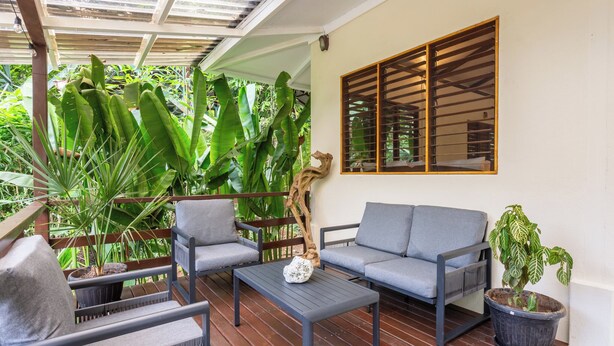 Un espacio de estar al aire libre cubierto con un sofá, sillas y una mesa de centro, rodeado de grandes plantas verdes y una pérgola de madera.