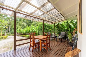 Una terraza de madera con mesa y sillas, rodeada de exuberante vegetación y un espacio cubierto con techo metálico.