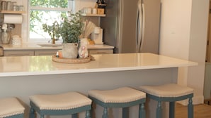 A kitchen with a large island, bar stools, a refrigerator, and pendant lights. - Coastal Getaway / 2BR/2BA Cottage + Bonus Room near Downtown & Cumberland Island (St. Marys)