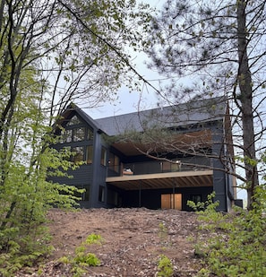 A modern two-story house with a balcony, surrounded by trees and a wooded area.