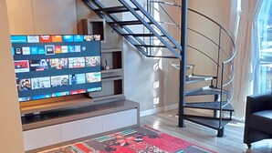 Una sala de estar moderna con una escalera, un televisor de pantalla plana mostrando varios canales y una alfombra colorida.
