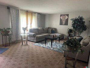 Een woonkamer met een bank, salontafel en vloerkleed. Er staan planten en hangt een schilderij aan de muren.