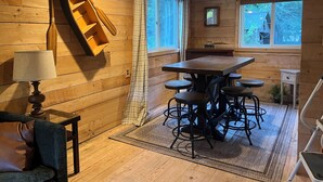 A cozy room with wooden walls, a dining table with stools, a lamp, and a window with curtains.