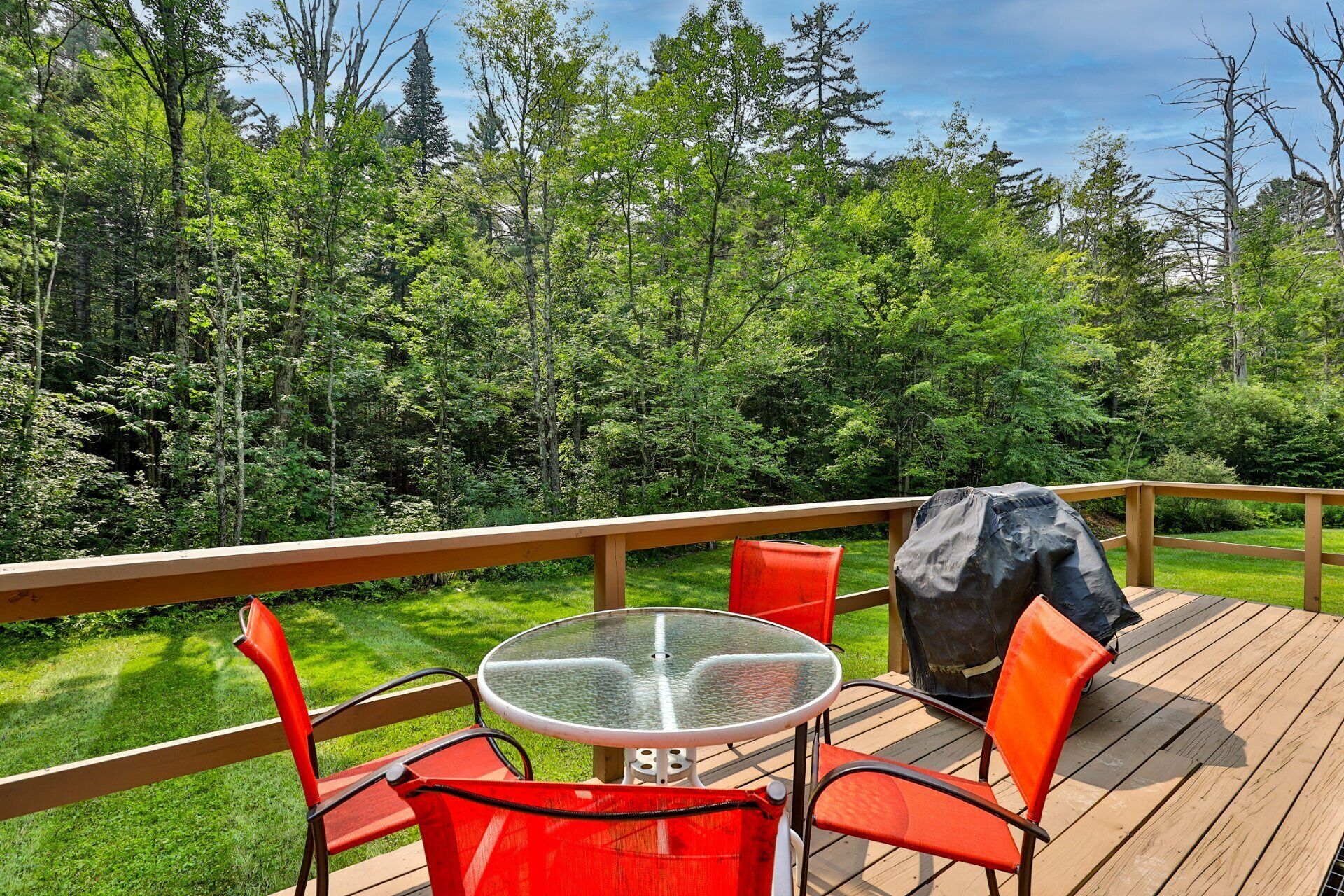 A deck with a glass-top table and red chairs, overlooking a forested area.