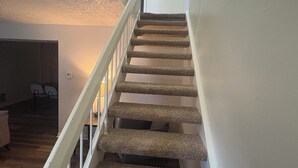 Un escalier recouvert de moquette, avec des rampes blanches, menant à une pièce éclairée par un luminaire au plafond.