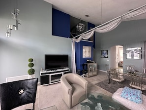 A modern living room with a flat-screen TV, a sofa, a glass coffee table, and a dining area with metal chairs.
