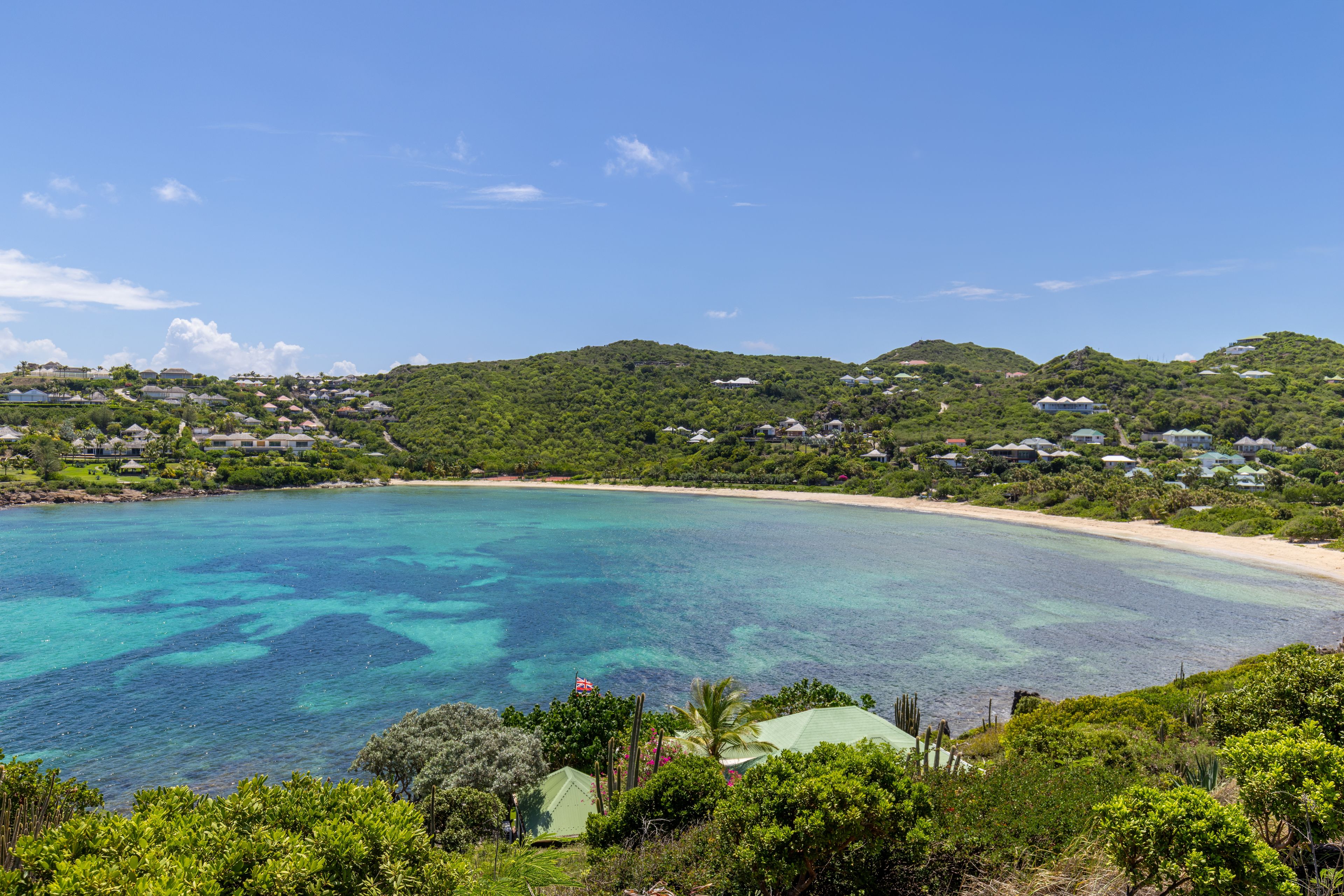 A coastal landscape with a clear blue sea, a sandy beach, and a hill with houses.