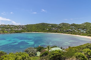 A coastal landscape with a clear blue sea, a sandy beach, and a hill with houses.