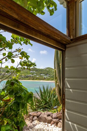 A view through a wooden-framed window overlooking a beach with houses on a hillside, a clear sky, and a cactus plant in the foreground.