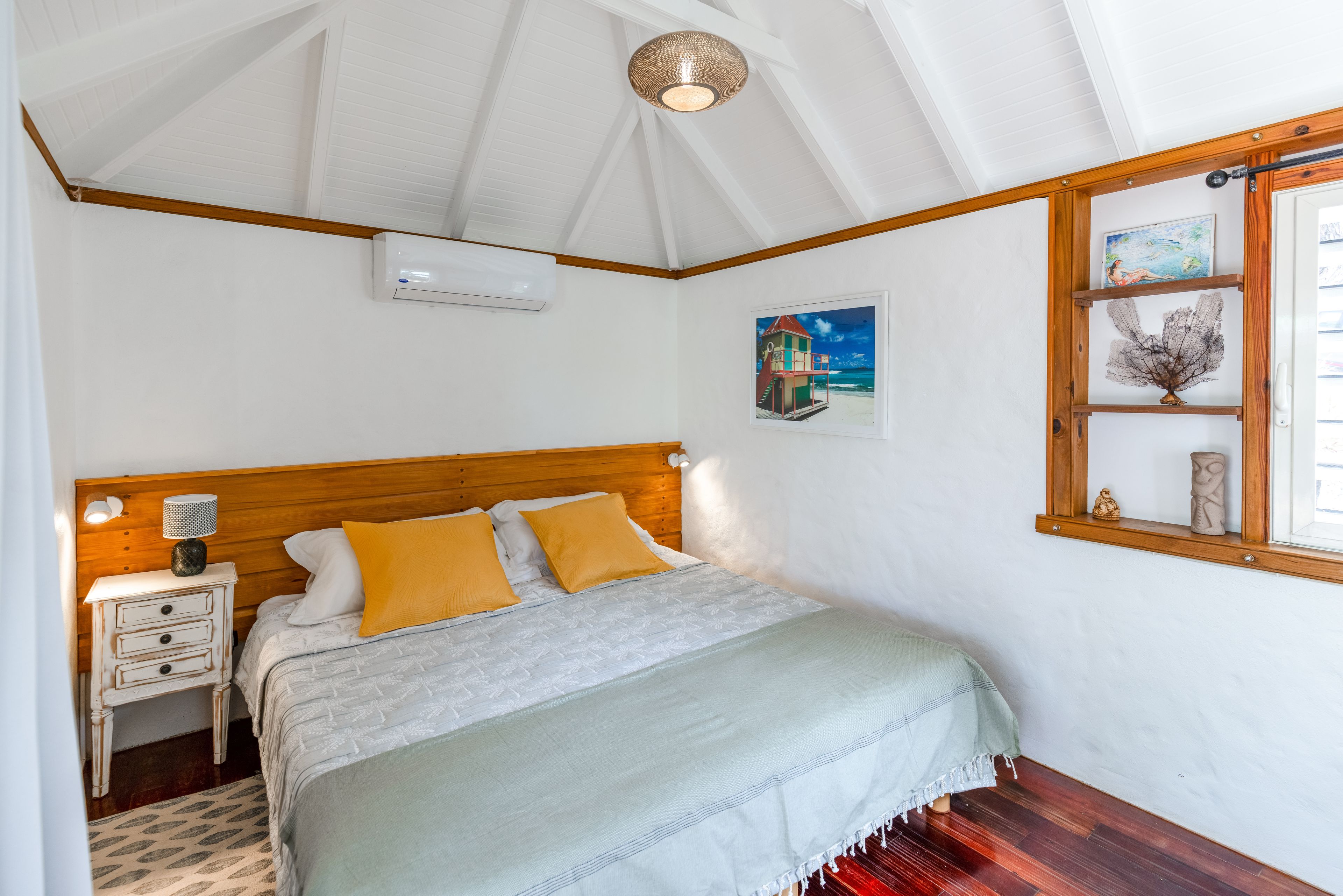 A bedroom with a wooden headboard, a bed with a white and green bedspread, a nightstand with a lamp, and a window with a view of the beach.