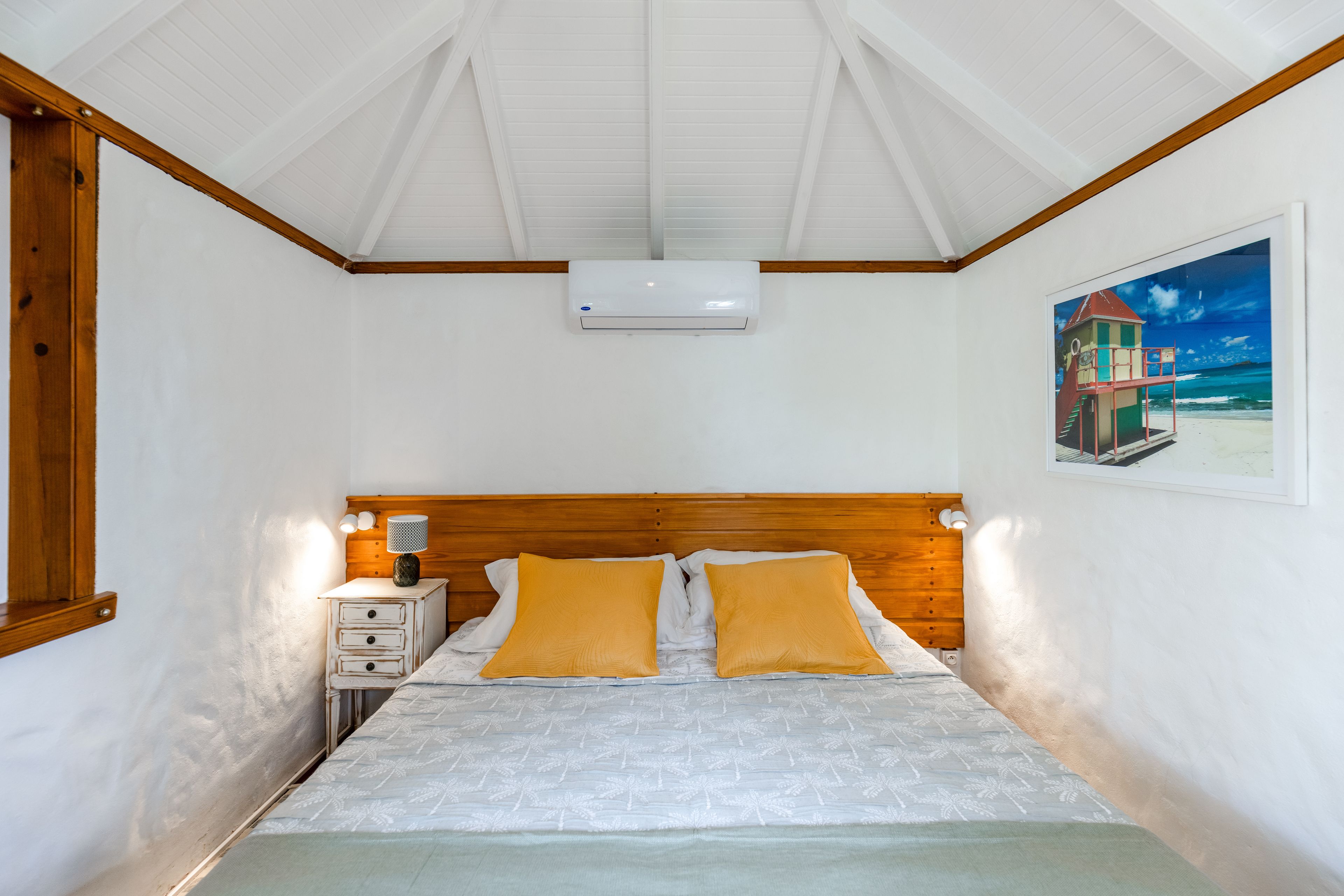 A bedroom with a bed, a nightstand, an air conditioning unit, and a painting on the wall.
