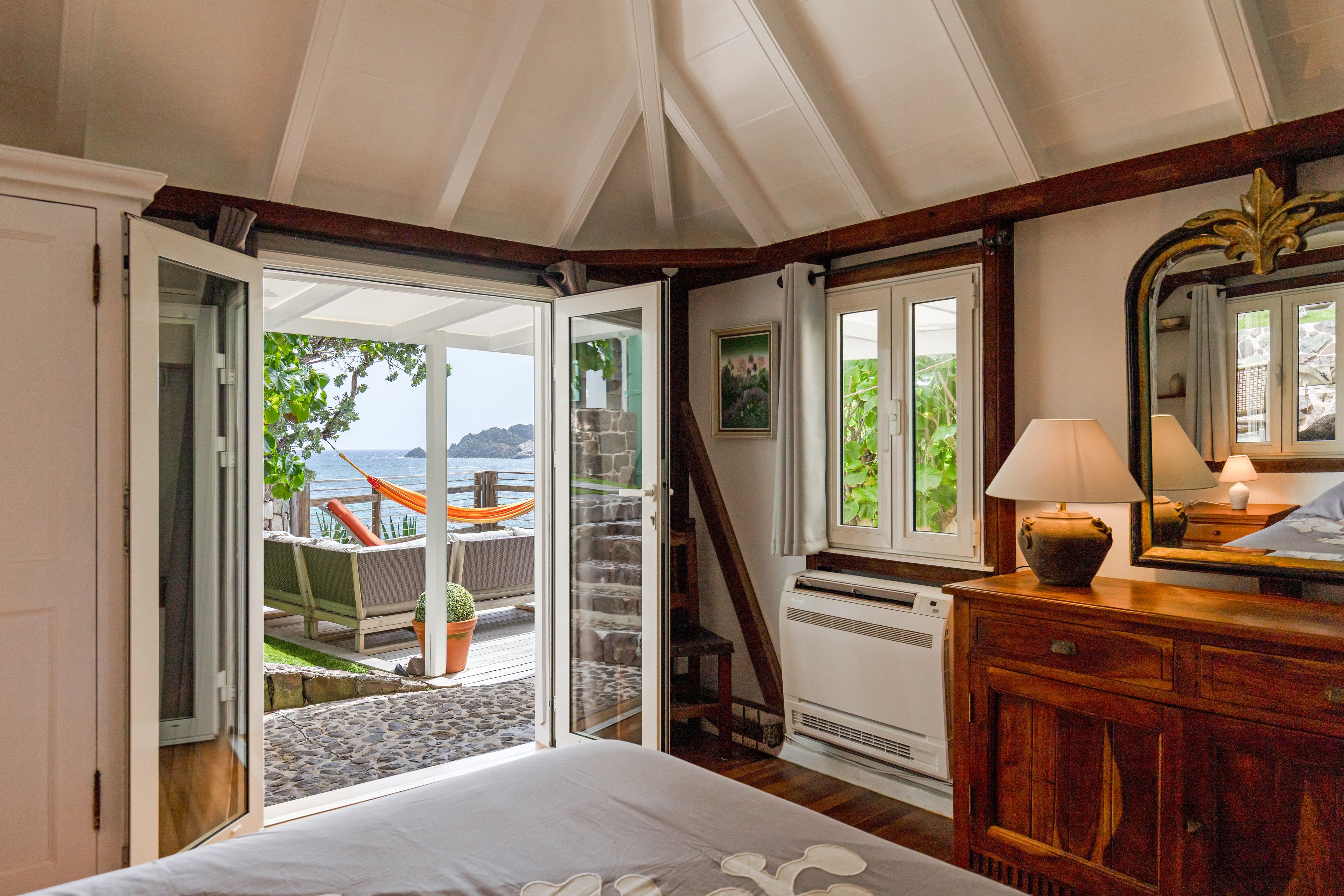 A bedroom with a large bed, a wooden dresser, a nightstand with a lamp, a window with a view of the outdoors, and a door leading to a balcony with a view of the sea.