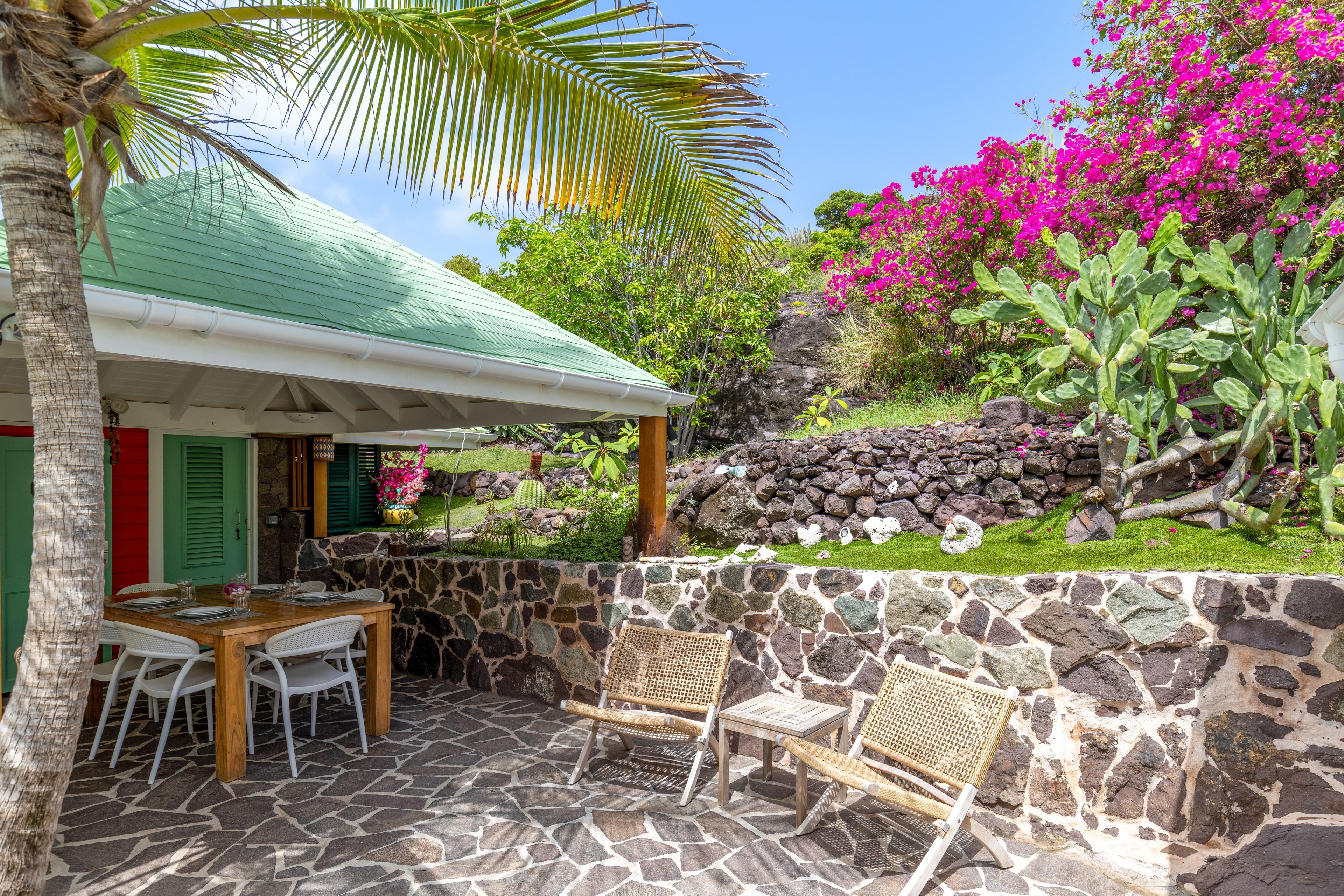 Un patio de piedra con una mesa y sillas, un muro de piedra y un jardín exuberante con flores rosadas y cactus.