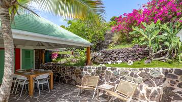 Un patio de piedra con una mesa y sillas, un muro de piedra y un jardín exuberante con flores rosadas y cactus.