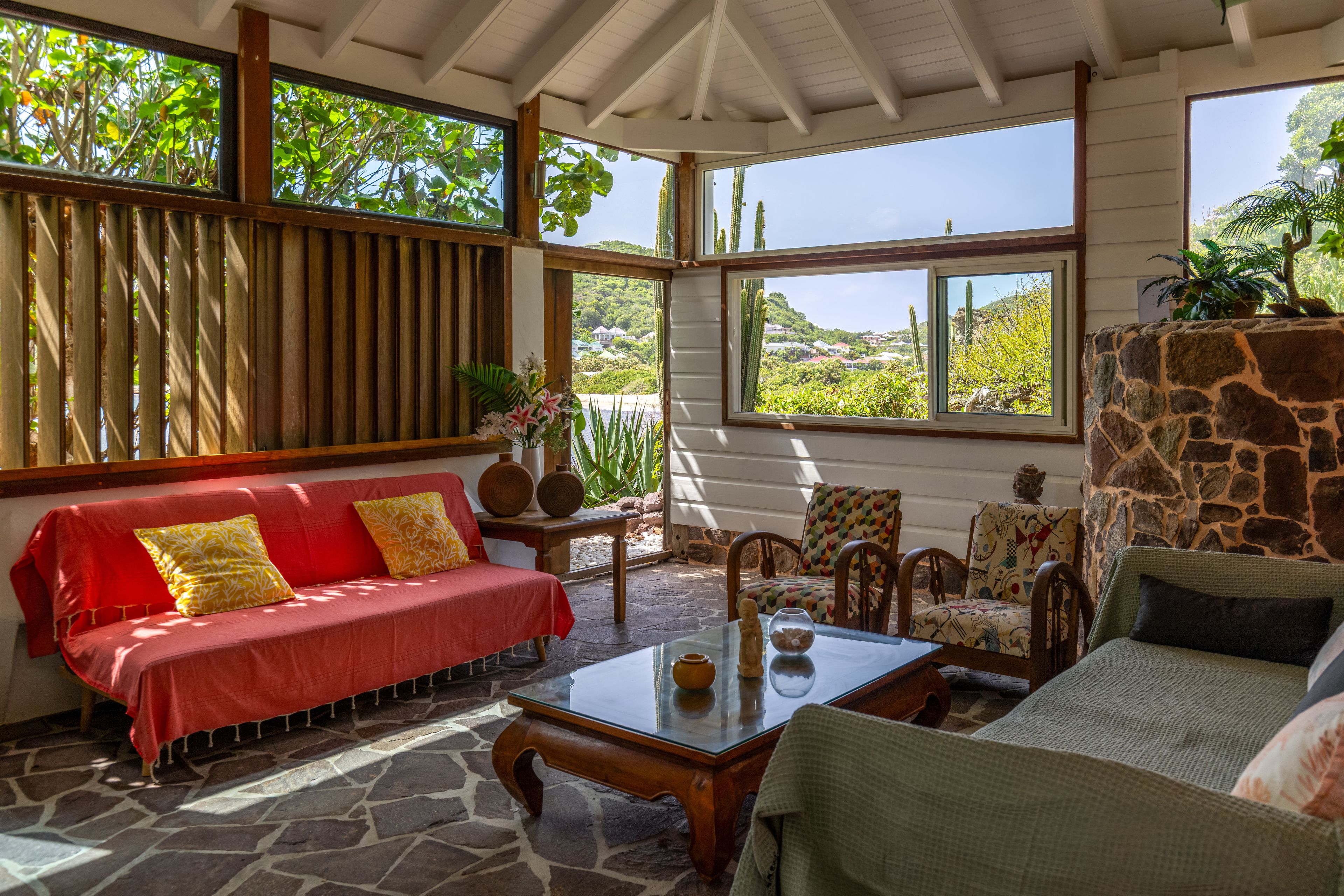 A sunlit room with a red sofa, patterned armchairs, and a stone wall.