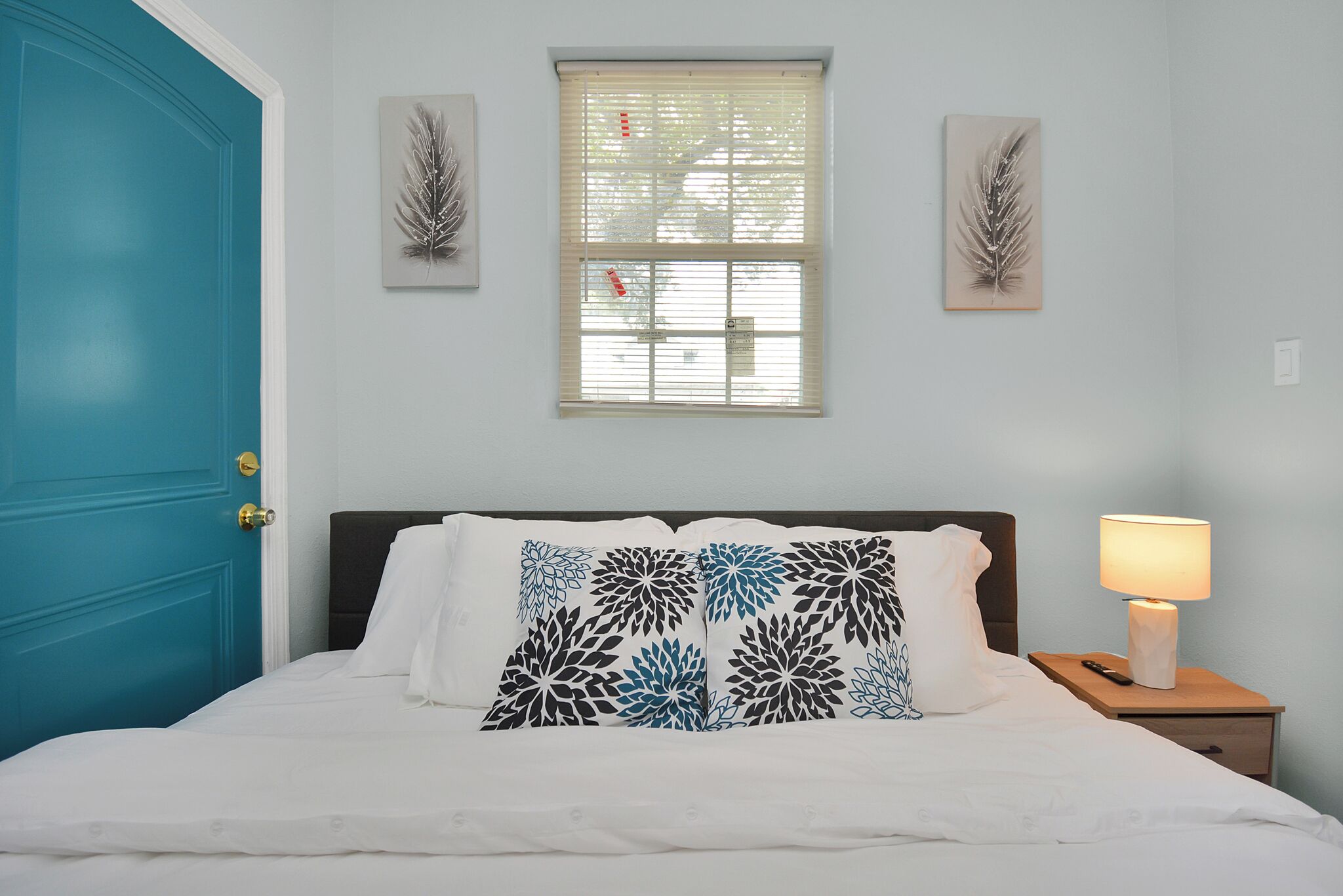 A bedroom with a blue door, a bed with white bedding and decorative pillows, a nightstand with a lamp, and two wall-mounted feather art pieces.